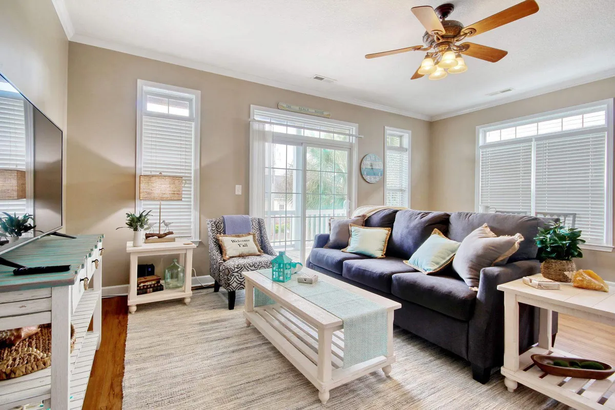 The image showcases a bright and inviting living room, featuring a plush sofa and coffee table set before large windows that fill the space with natural light. Stylish decorative elements create a warm, coastal atmosphere ideal for relaxation or gathering with loved ones.