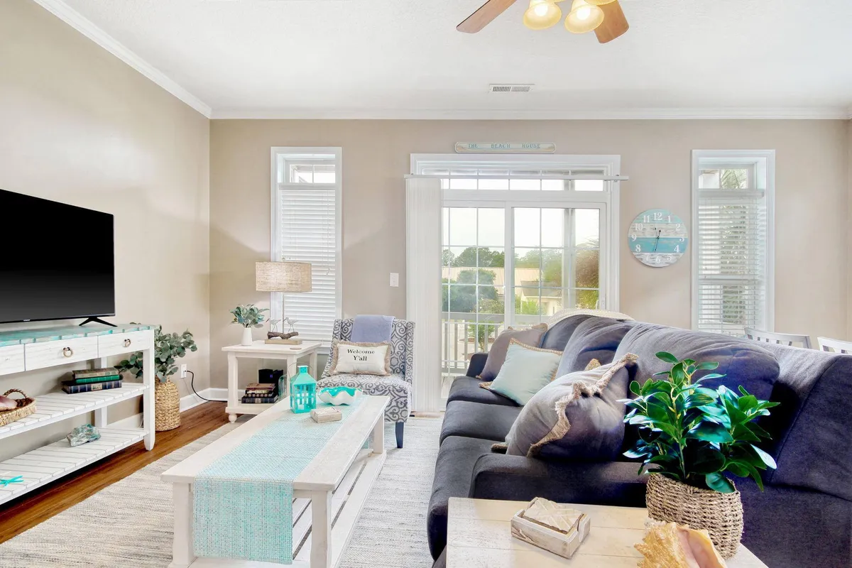 A bright and inviting upstairs living area featuring a comfortable sofa, a coffee table ready for board games or snacks, a television for movie nights, lush green plants, and large windows that fill the room with natural light. The space feels open and cozy—perfect for relaxing, reading, or gathering as a group.