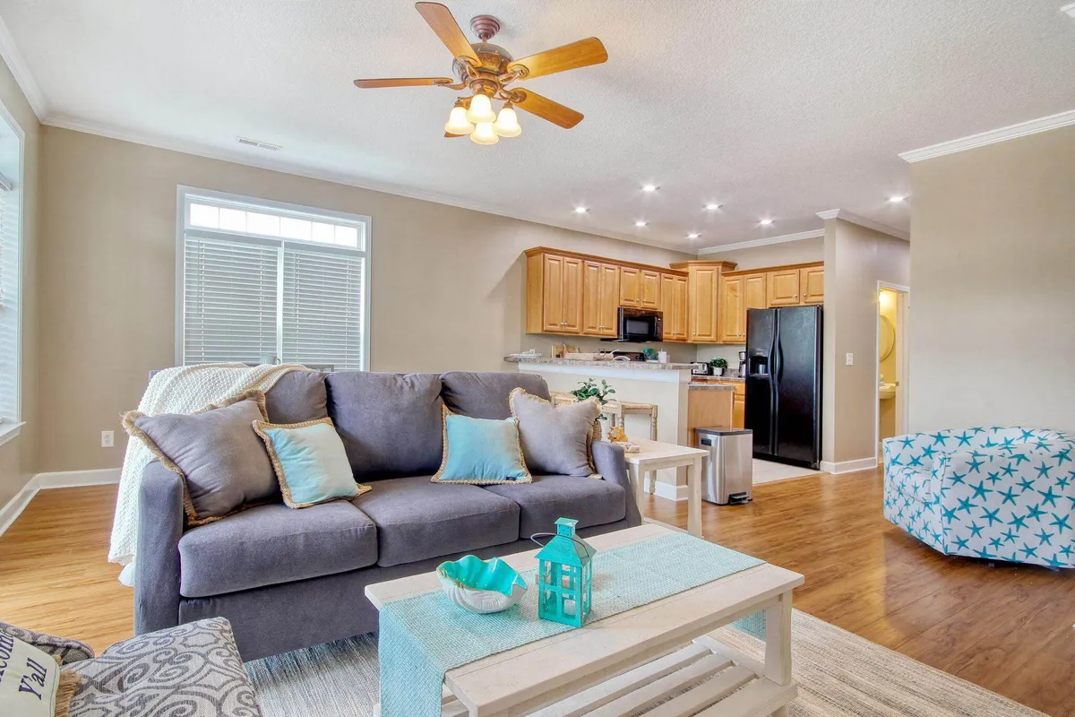 A bright and open living area featuring a comfortable sofa with decorative cushions, a coffee table with charming decor, and a glimpse of the kitchen in the background. The space is styled with coastal décor and bathed in natural light, creating an inviting place for gathering and relaxation.