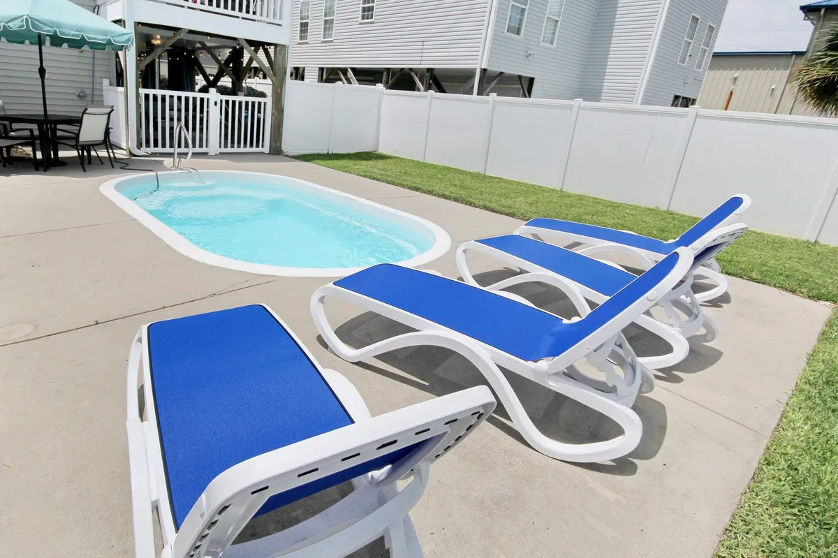 An exterior view of a swimming pool surrounded by lounge chairs and a fenced yard, signaling a leisure-focused outdoor space for sunbathing, swimming, or outdoor dining. The setup hints at relaxation and fun, with room for gatherings and barbecues.