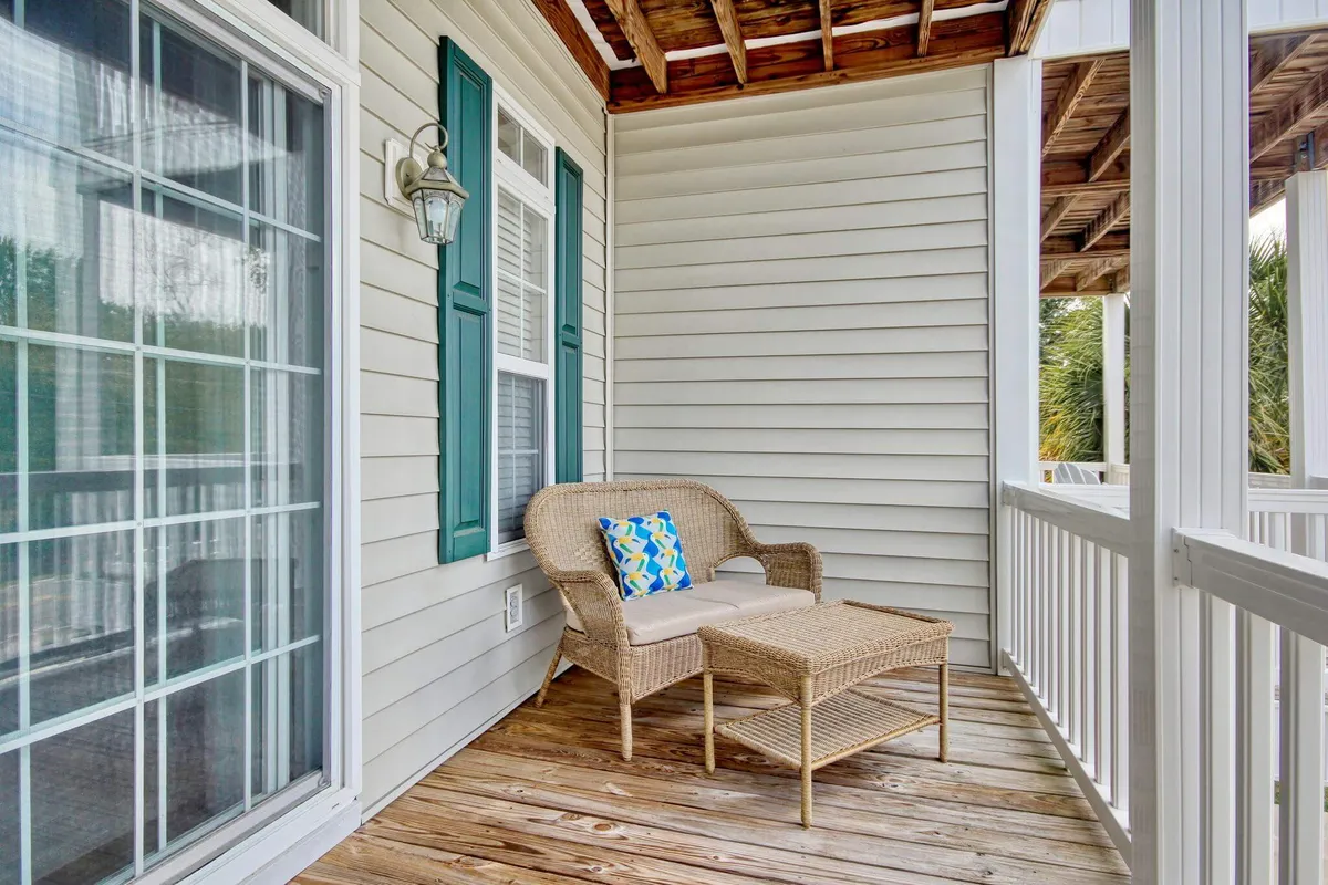 A serene porch scene with a wicker chair, colorful pillow, wooden flooring, and green shutters. The inviting setup highlights an ideal spot for enjoying morning coffee, peaceful afternoons, or evening stargazing in a coastal setting.