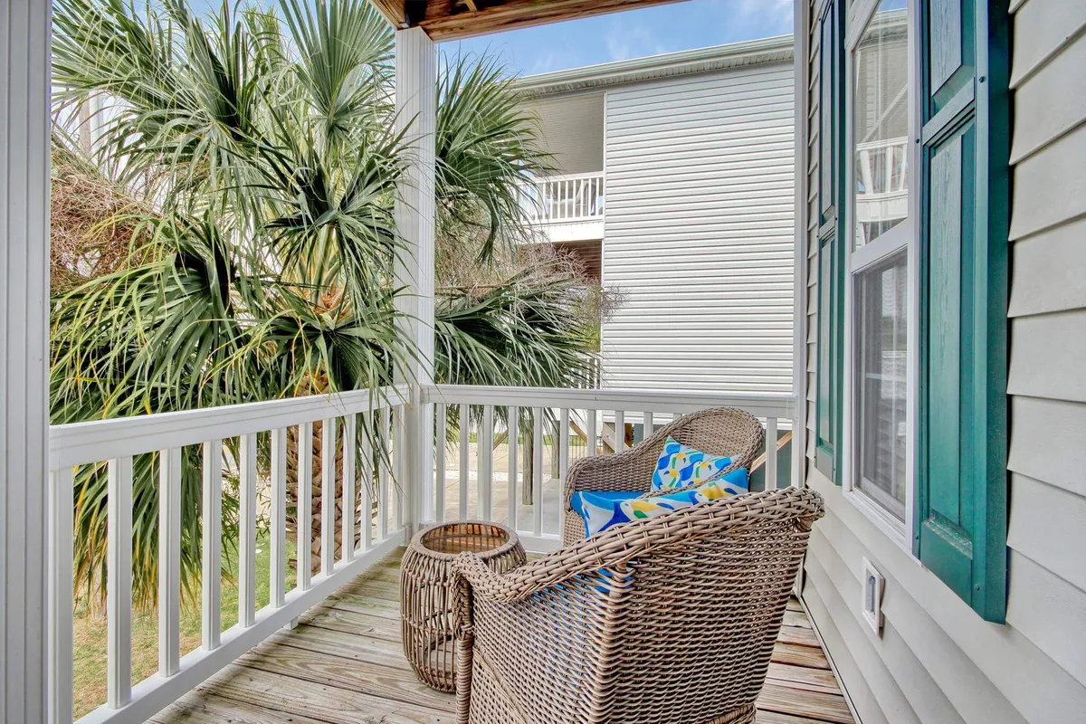 An upper porch scene with palm trees in the background, wicker chairs with colorful throw pillows, and a wooden deck. The peaceful ambiance suggests a spot for sipping morning coffee, reading, or simply enjoying the sea breeze.