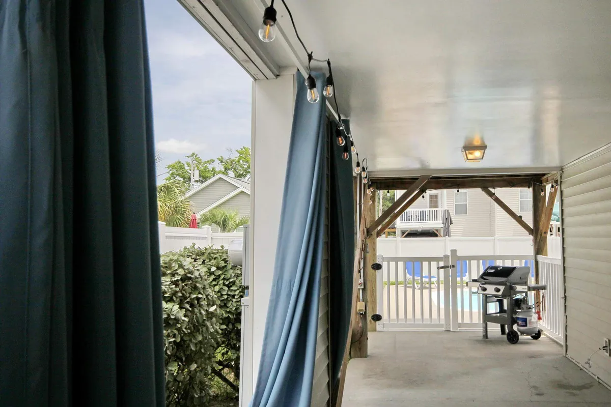 A covered outdoor area featuring an outdoor grill, pool area views, string lights, and comfortable seating. The setup suggests a casual, family-friendly space ideal for barbecues, socializing, and relaxing after a day at the beach.