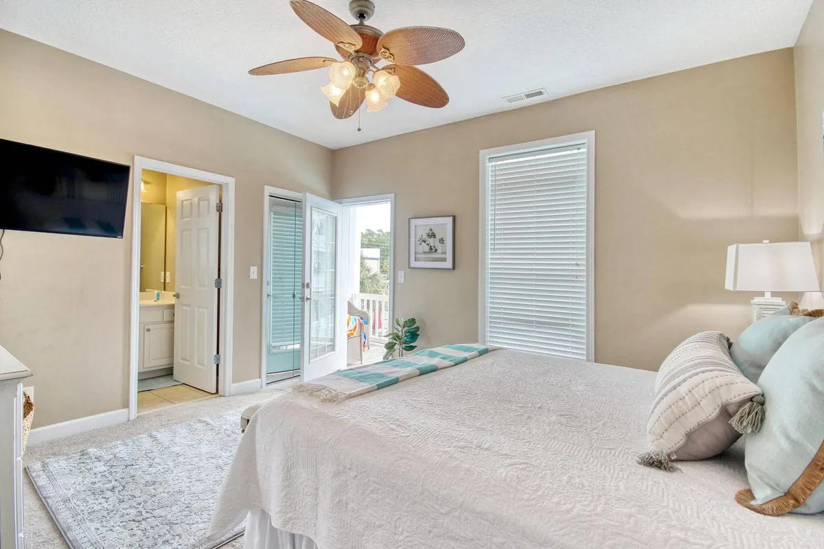 A serene bedroom featuring a large bed with crisp bedding, a ceiling fan overhead, tasteful artwork on the wall, and doors opening to a balcony. Natural light floods the room, hinting at easy access to a private outdoor space.