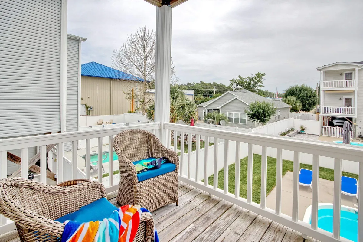 This exterior image features wicker chairs with colorful towels arranged on a deck overlooking the pool area and green lawn. The inviting seating area is positioned to enjoy the view, suggesting a perfect spot for socializing, relaxing, or sharing meals outdoors while soaking in the sunset or ocean breeze.