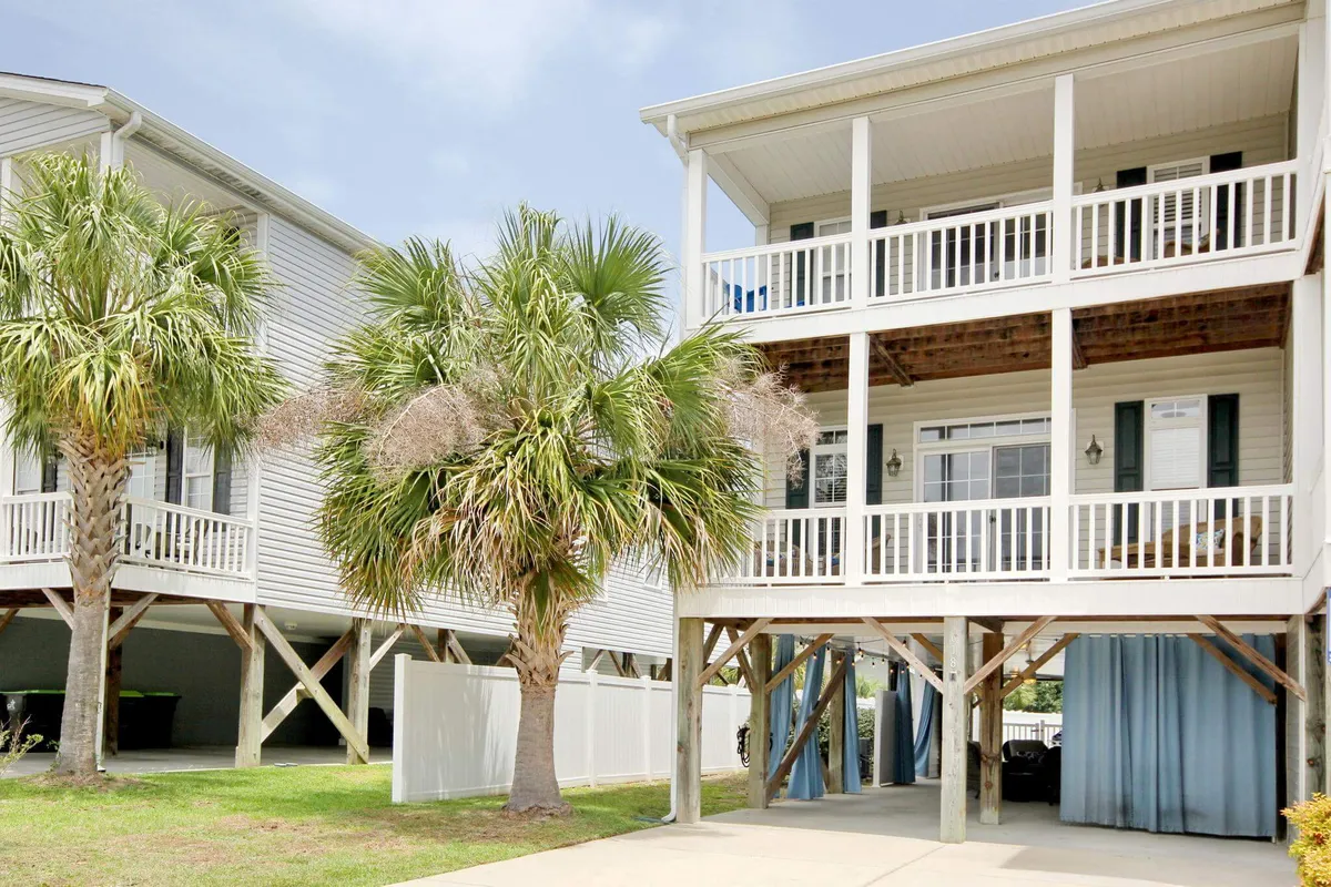 This exterior scene highlights the balcony surrounded by palm trees, exposed beams, and a cozy gathering space under the house. The image captures the relaxed, coastal environment, with hints of the neighborhood’s charm and natural beauty. It visually connects the home to the surrounding beach area, reinforcing the idea of seamless indoor-outdoor living and easy access to local attractions.